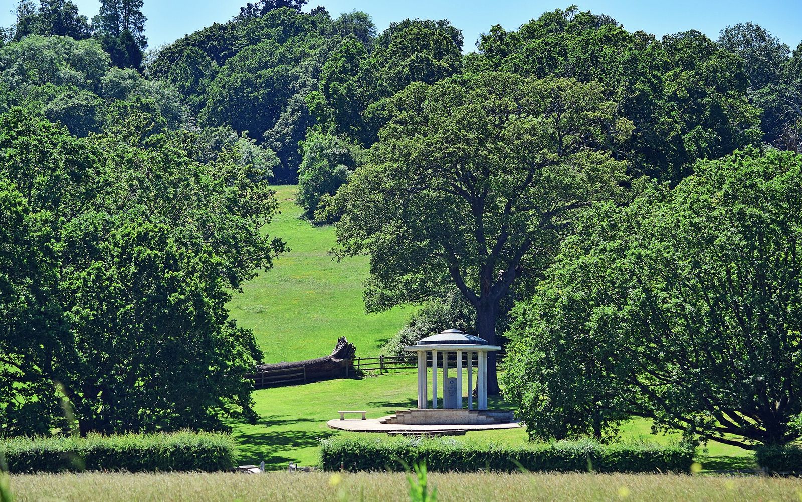 A small white classical rotunda stands in a green meadow at the foot of a wooded hill: the Magna Carta Memorial at Runnymede, built by the American Bar Association in 1957.