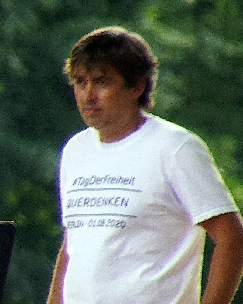 Michael Ballweg, in a white Querdenken T-shirt dated Berlin 01.08.2020, standing on a stage in front of trees.