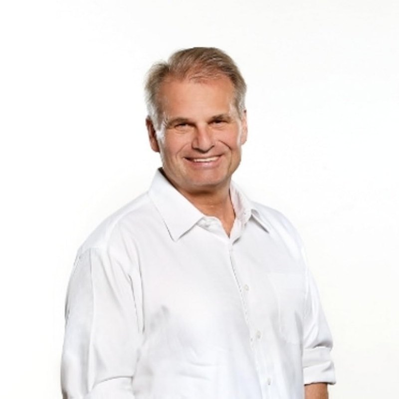 Portrait of Reiner Fuellmich, smiling, in a white open-collared shirt against a plain white background.