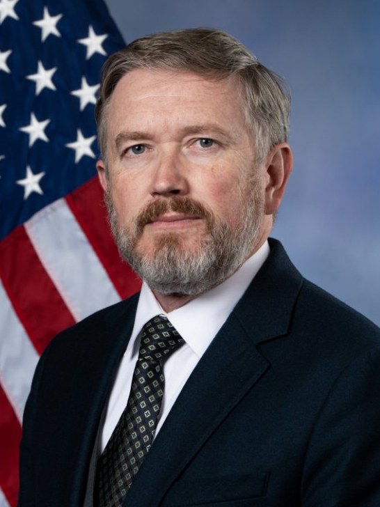 Official 119th Congress portrait of Rep. Thomas Massie of Kentucky, with a gray beard, in a navy suit and patterned tie, in front of an American flag.