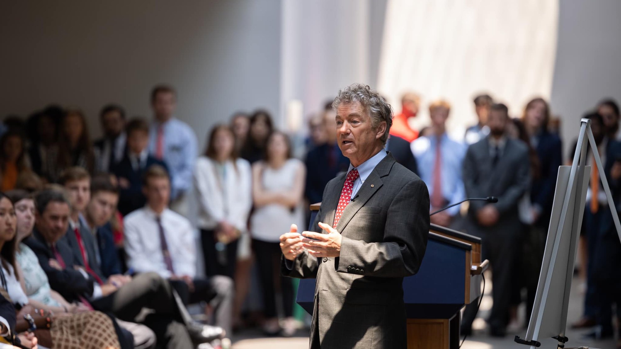 Senator Rand Paul speaking at a lectern before a standing audience of young staff and visitors in a Senate office building.